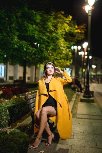 A vertical shot of a young blonde female model in a fashionable yellow coat posing on a bench