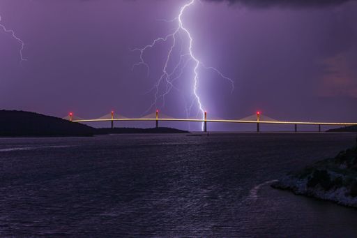 A picturesque view of a bridge over a body of water, illuminated by a spectacular display of lightning in the background