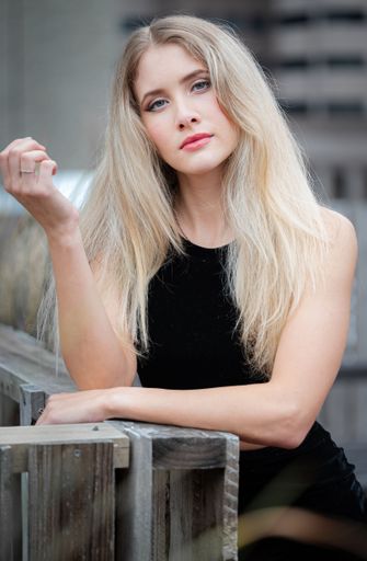 A blonde girl wearing a black evening dress leaning on a wooden railing in an urban setting