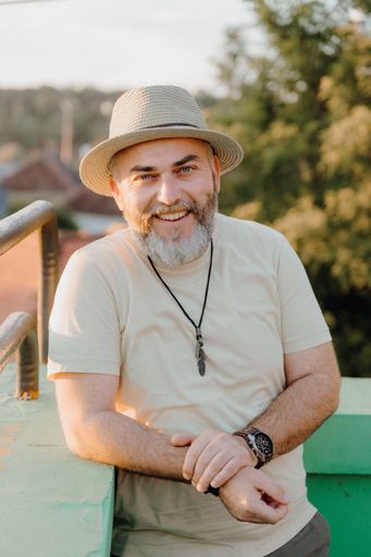 A mature man wearing a hat is sitting in a relaxed pose, facing the camera and smiling