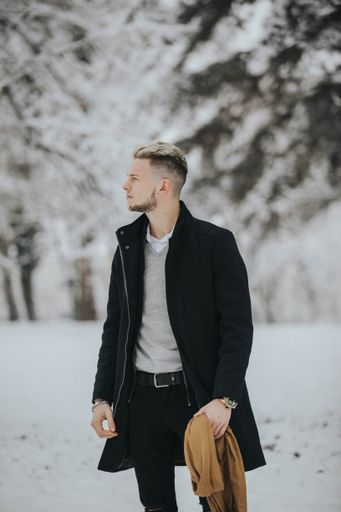 A handsome young man in a black winter outfit walking in a snowy forest path holding his brown scarf