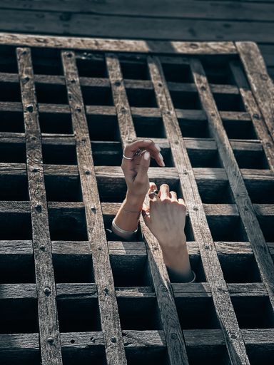 A female hand wearing silver rings reaches out from a wooden prison gate, symbolizing a longing for freedom