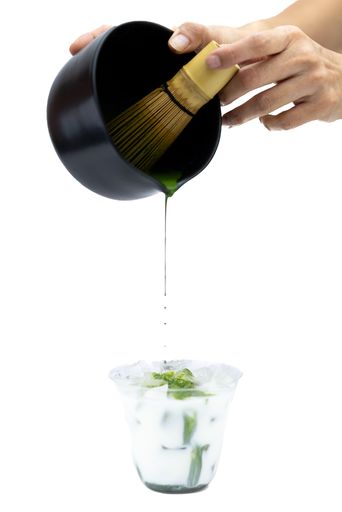 A hand pouring matcha into cup with ice and milk on a white background - process of making matcha tea