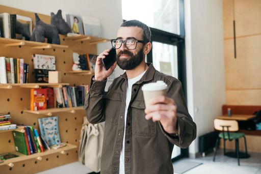 A young Caucasian man in his 20s is standing outside, talking on his phone while holding a cup of coffee in his left hand