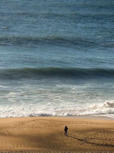 A silhouette of a person standing on a sandy beach looking at the splashing waves at sunset