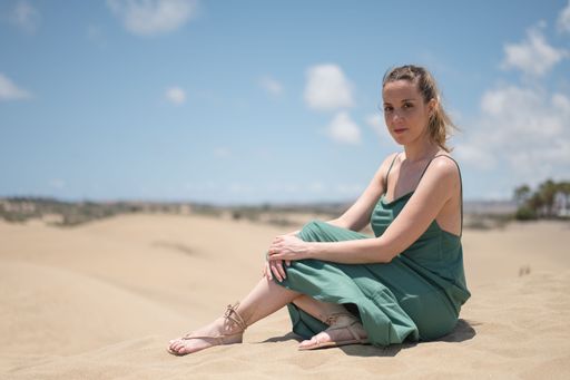Blonde young girl in green dress sitting on sand of beach dunes