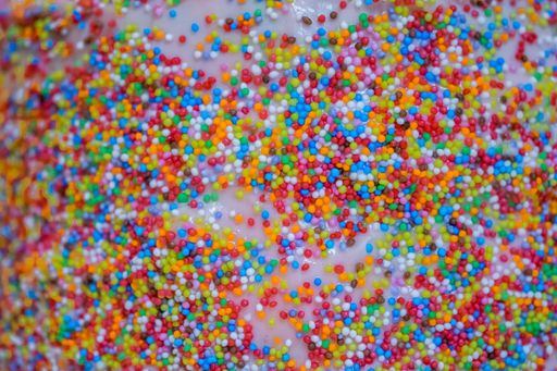 A pink-themed birthday cake, decorated with pink and white marshmallows and sweets and covered with colorful small sprinkles on the sides.