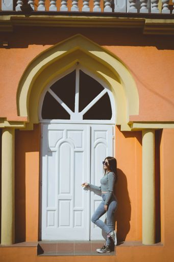 An adult multiracial woman wearing jeans with a striped shirt and leaning on a residential building