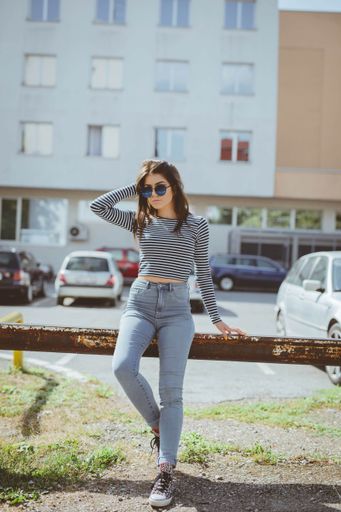 A vertical shot of a beautiful Caucasian woman wearing a striped shirt and jeans, posing outdoors, leaning on a railin