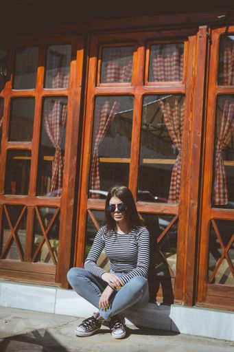A Bosnian brunette girl in a cool summer outfit sitting on a sidewalk in front of a wooden window wall