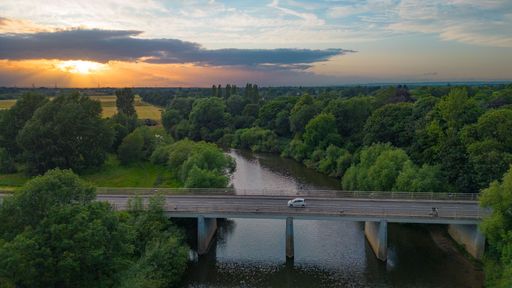 A white car is driving across a picturesque bridge over a tranquil river, surrounded by lush green trees and the sun setting behind clouds.