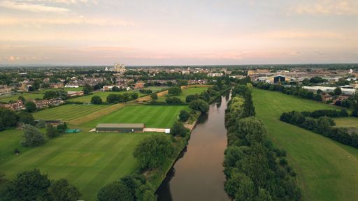 An aerial view of a river flowing through a lush green field with rural houses at sunset