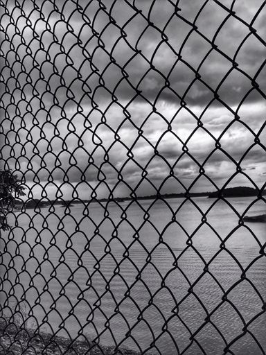 A chain-link fence against the background of a cloudy sky and tranquil lake.