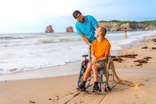 A  closeup of a man  in an orange T-shirt in a wheelchair with his friend smiling on a sandy beach