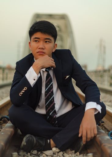 A closeup shot of a young Asian man in a suit sitting in the middle of a railway with chin on hand