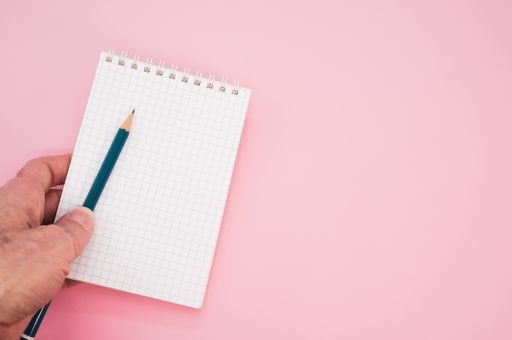 A closeup shot of a hand holding a pencil with a notebook on a pink background