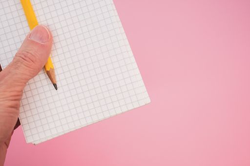 A closeup shot of a hand holding a pencil with a notebook on a pink background
