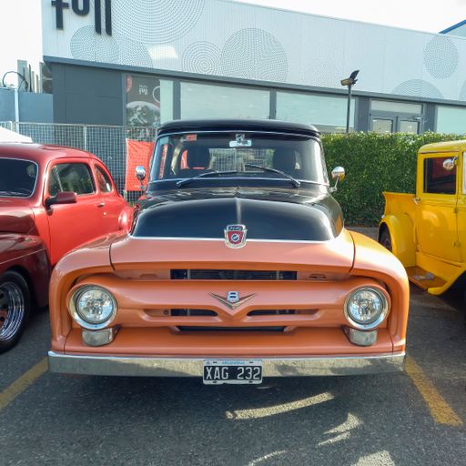 An old black and orange 1956 Ford F 100 V8 pickup truck in a parking lot at a classic car show
