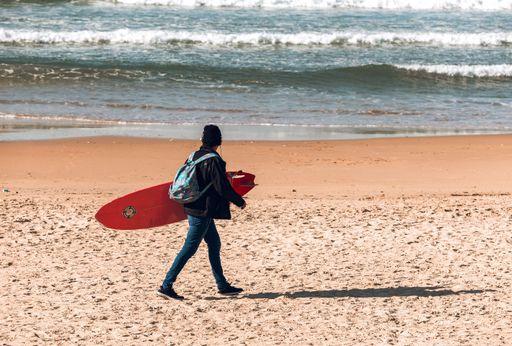 A young man walking along a sunny beach carrying a red surfboard