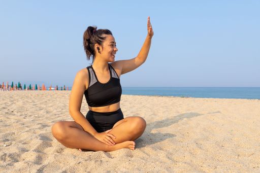 A young latina female in athleisure sitting and waving a on a beach