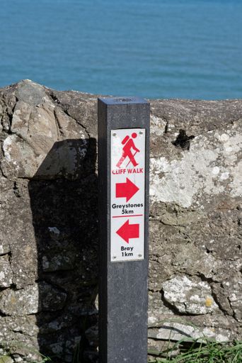 Vertical shot of the sign with distances to Greystones and Bray at the cliff walk trail in Co. Wicklow, Ireland. Stone wall and sea in the background.