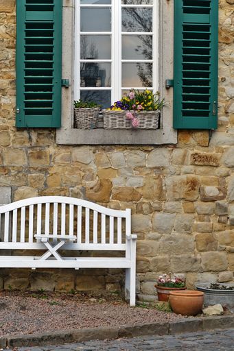 A vertical shot of a stone building facade with flowers, a bench, and green window shutters