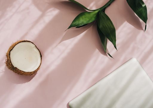 A top view shot of a pink surface, a laptop on it, green plant leaves, and half of a coconut