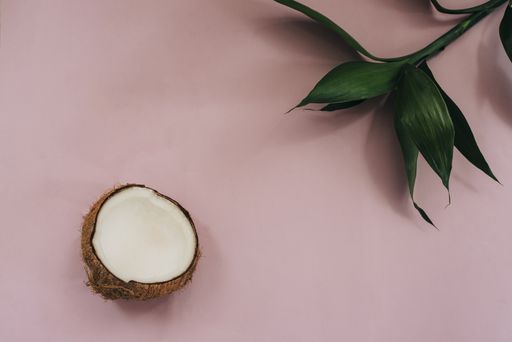 A top view shot of a pink surface, green plant leaves, and half of a coconut