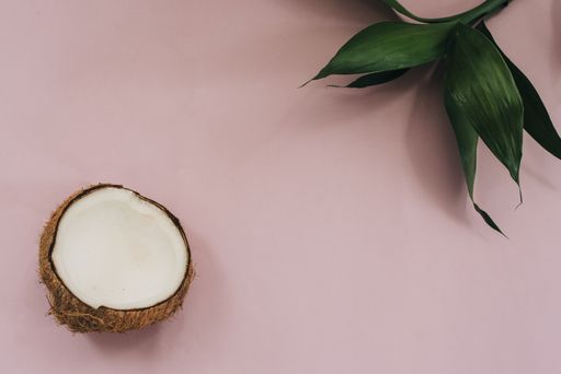 A top view shot of a pink surface, green plant leaves, and half of a coconut