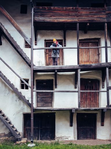 An elderly man wearing a hard hat is standing on the balcony of a residential building, looking out into the distance