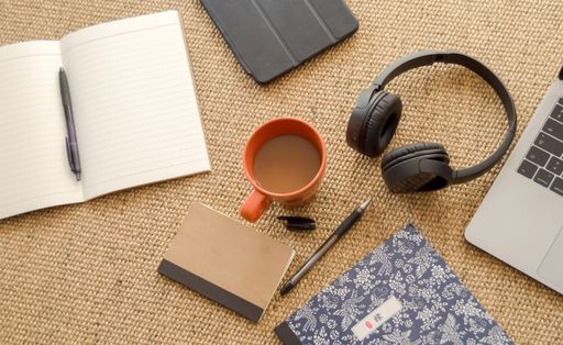 A cup of coffee with notebooks, ballpens, headphones, and laptop on brown burlap textile