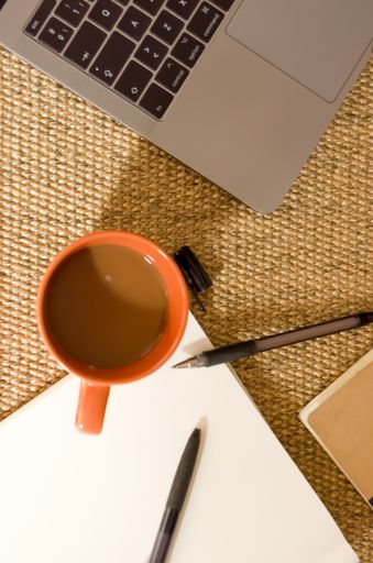 A cup of coffee with notebooks, ballpens, and laptop on brown burlap textile