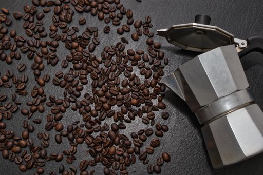 A top view of the Moka pot on the black surface covered with scattered roasted coffee beans