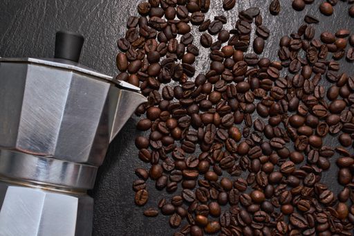 A top view of the Moka pot on the black surface covered with scattered roasted coffee beans