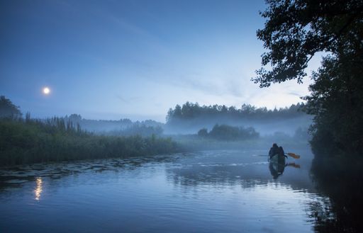 Hikers rowing an canoe in the summer night river, surrounded by fog clad floodplain and under full moon in the sky on the Halliste river in Soomaa NP