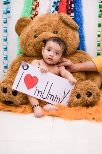 A vertical shot of a cute happy Indian female baby sitting on a teddy bear holding a poster for mom