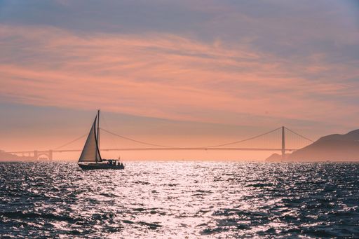 A beautiful shot of waves reflecting the sky with a sailboat sailing with a bridge in the distance at San Francisco