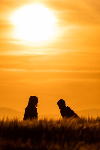 A vertical shot of the silhouette of a couple sitting on top of a hill at sunset