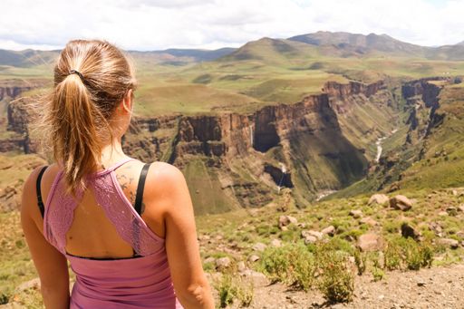 A girl standing on rocks surrounded by hills covered in greenery under the sunlight