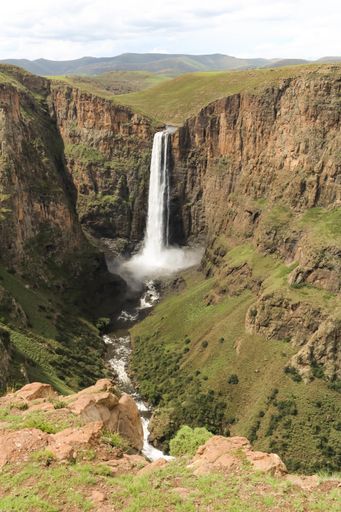 A vertical picture of a waterfall surrounded by rocks covered in greenery under the sunlight
