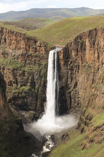 A vertical picture of a waterfall surrounded by rocks covered in greenery under the sunlight