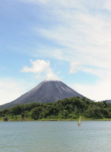 A vertical shot of the Arenal volcano peak and a lake with forests on the shore in the foreground