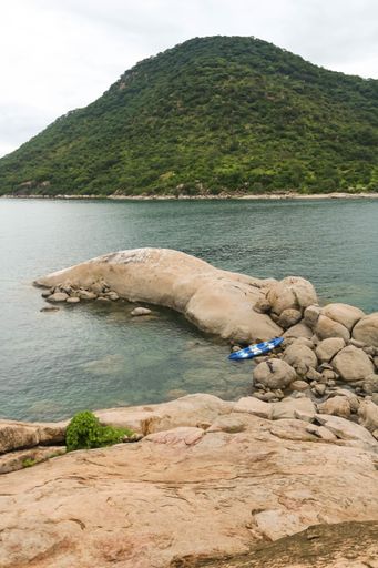 A vertical picture of the sea surrounded by rocks and hills covered in greenery under a cloudy sky