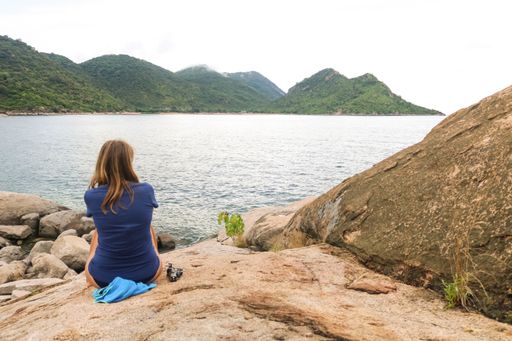 A woman sitting on rocks surrounded by the sea and hills covered in greenery