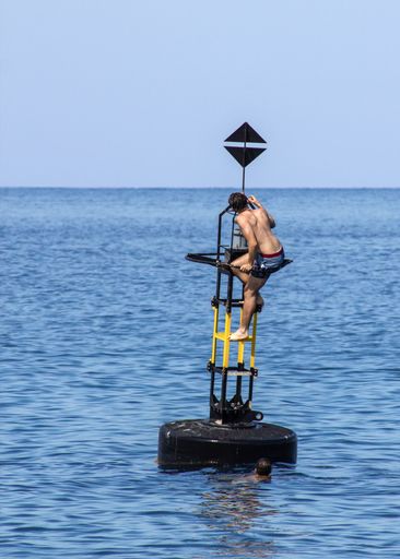 Some teenage boys scaling a tall white buoy in the sparkling blue waters of Marsalforn, Gozo, Malta