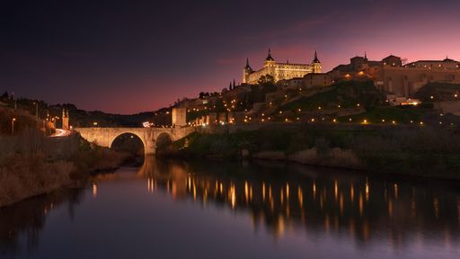 A beautiful shot of a classical architectural building with glowing lights and reflective river with a scenery of sunset in the background