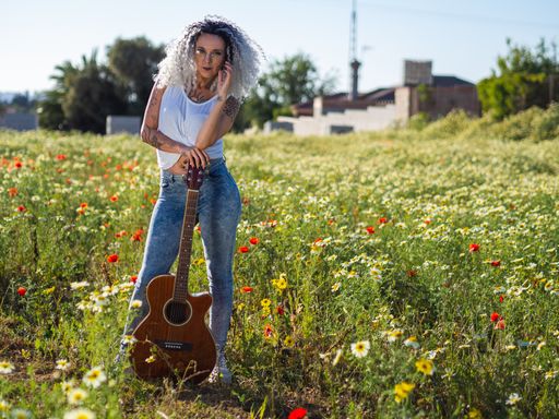 A smiling cute Spanish woman with white curly hair and cool tattoos posing in a meadow with her guitar
