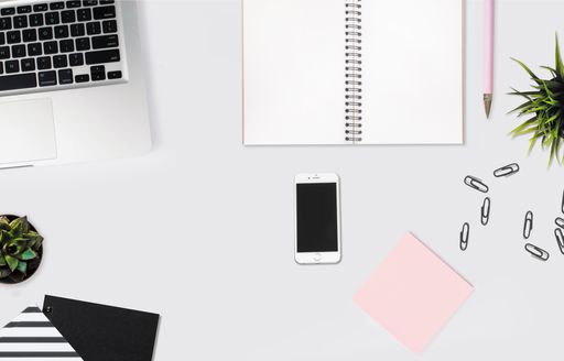 An overhead shot of a smartphone on a white desk with a notebook, pink sticky notes and paper clips