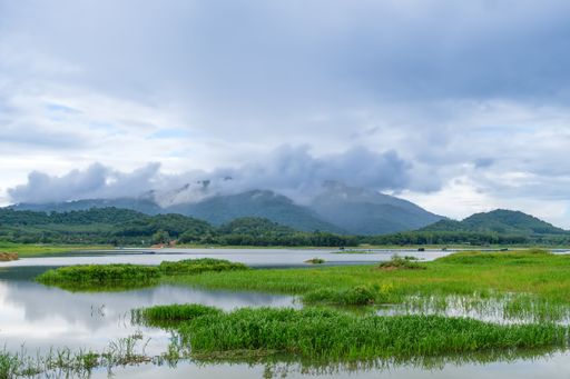A scenic landscape of majestic mountains partially obscured by cloudy skies, framed by a tranquil lake and lush meadows below