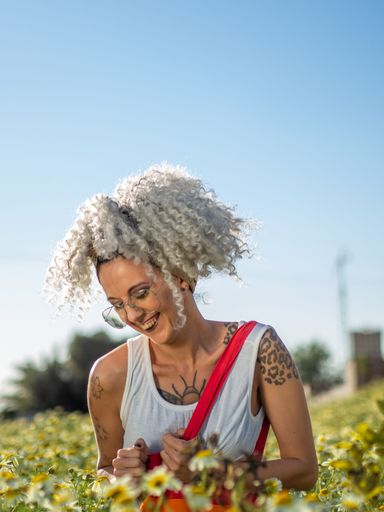 A shallow focus of a young woman with white curly hair and tattoos holding a tote bag with a pride flag print in a field